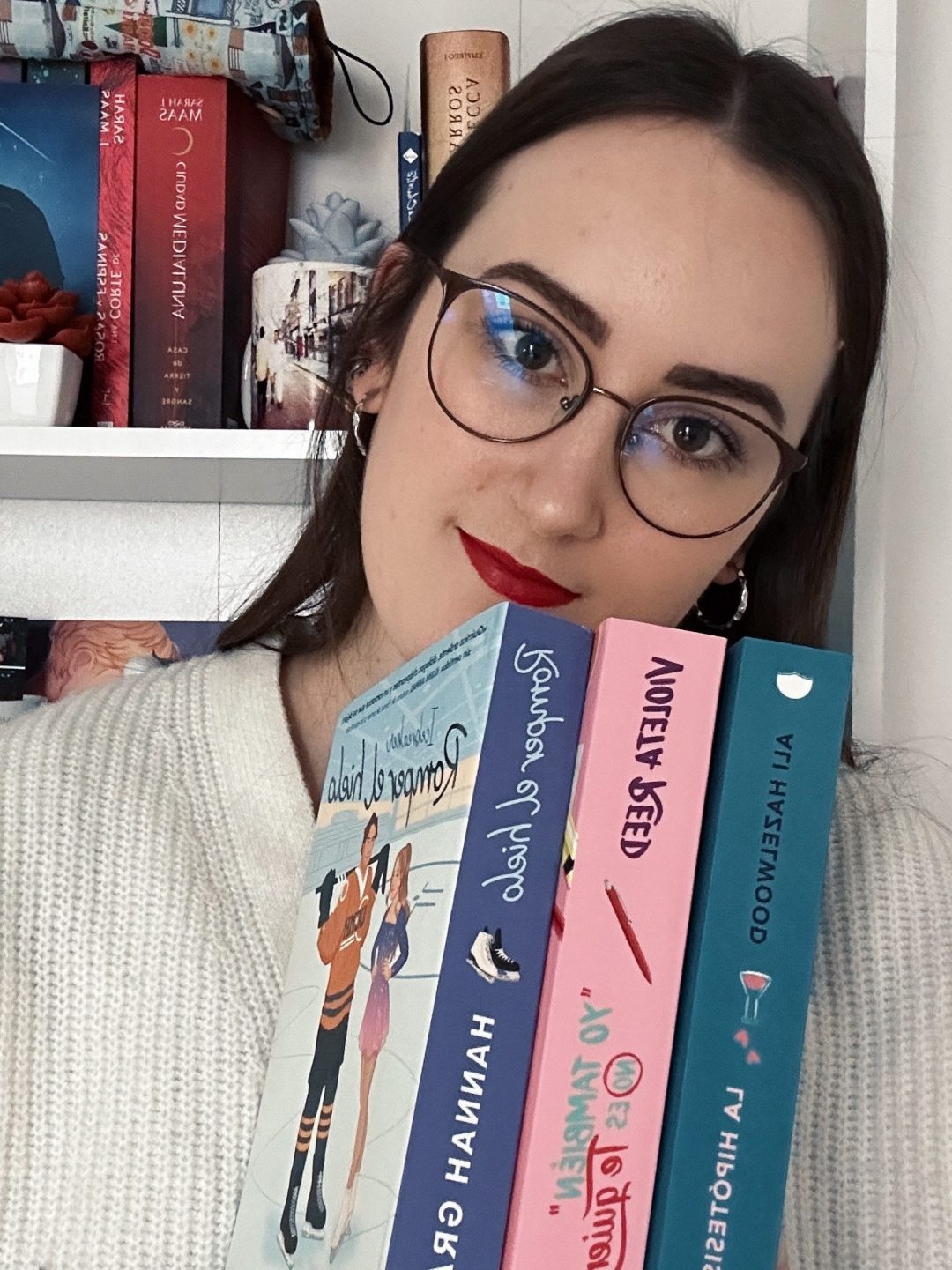 Una persona joven con gafas sostiene varios libros frente a una estantería decorativa. Los libros tienen cubiertas coloridas y el fondo muestra más libros y decoraciones.