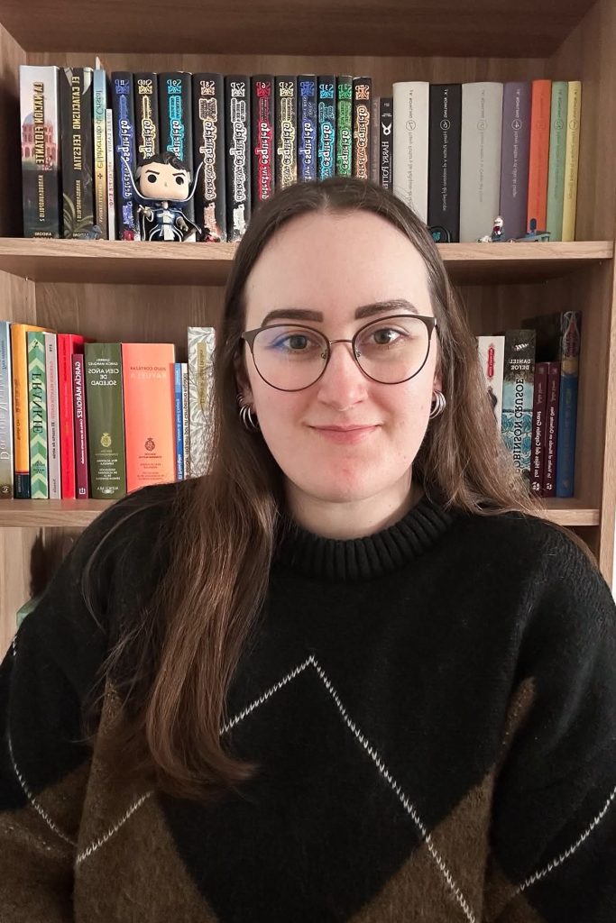 Retrato de una persona con suéter negro de patrones, sonriendo frente a estanterías llenas de libros.